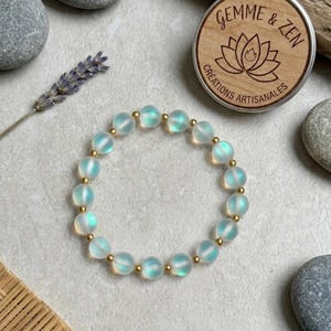 Peut inclure: Un bracelet composé de perles en verre translucide irisé aux reflets bleutés alternées avec de petites perles intercalaires métalliques dorées, disposées en cercle sur une surface neutre près d'un couvercle de boîte en bois.