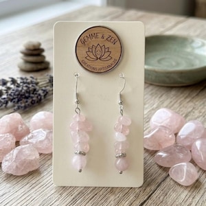 Peut inclure: Boucles d'oreilles pendantes composées de perles de quartz rose. Elles sont présentées sur une carte crème avec un logo en bois "Gemme & Zen". Des pierres de quartz rose sont dispersées autour.
