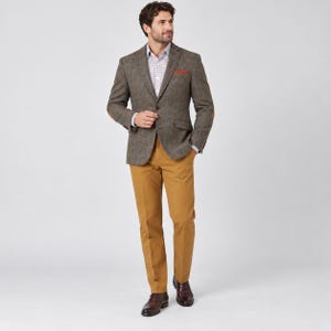 Pode incluir: Um blazer de tweed castanho com lapela entalhada e fecho de um botão, usado sobre uma camisa de botões de cor clara com padrão e um pequeno lenço de bolso laranja. O conjunto inclui calças amarelo-mostarda e sapatos de couro castanhos.