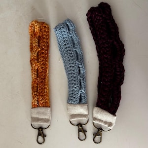 May include: Three wristlet keychains featuring textured crochet fabric in orange, light blue, and dark purple. Each strap is attached to a beige fabric connector and a bronze-toned metal lobster clasp, providing a functional way to carry keys.
