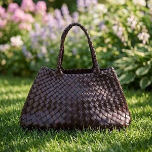 Puede incluir: Un bolso tote tejido marrón oscuro con un asa trenzada. El bolso está hecho de un material tejido y tiene forma trapezoidal. El bolso está sobre hierba verde con flores en el fondo.