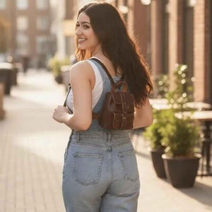 Könnte beinhalten: Ein brauner Lederrucksack mit mehreren Fächern wird über den Schultern getragen. Die Person trägt ein weißes Tanktop und eine Jeans-Latzhose und geht auf einer gepflasterten Straße. Im Hintergrund sind Gebäude und Grün zu sehen.