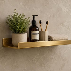 Puede incluir: Un estante de pared de metal dorado cepillado que sostiene una pequeña planta verde en maceta, una botella de vidrio ámbar oscuro con bomba y etiqueta blanca, un vaso beige estriado con dos cepillos de dientes de madera y una toalla beige.