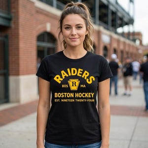 May include: A black short-sleeved t-shirt featuring yellow text that reads RAIDERS, BOS R MA, BOSTON HOCKEY, and EST. NINETEEN TWENTY-FOUR. The design includes a central yellow emblem with the letter R inside a keystone shape on the chest.