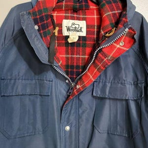 May include: A navy blue jacket featuring a red, green, and white plaid lining. The jacket includes a hood, a front zipper closure, and two large front pockets. A white label with the text Woolrich is stitched inside the collar area.