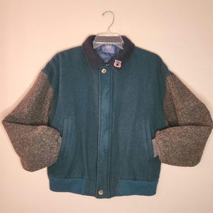 May include: A varsity-style jacket featuring a teal wool body and heathered grey sleeves. The jacket has a dark navy blue collar, a front button closure, and a small embroidered patch on the collar. It includes ribbed cuffs and a ribbed hem.