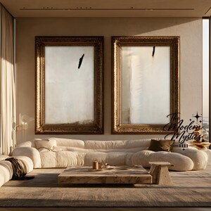 May include: Two large vertical rectangular wall art pieces in ornate gold frames featuring abstract white and cream canvases with single black brushstrokes. The artwork is displayed above a long, low-profile cream-colored sofa in a room.