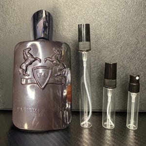 May include: A dark purple perfume bottle with a silver cap and a matching emblem, accompanied by three clear glass spray bottles with black caps. The perfume bottle has the text "PARFUMS de MARLY" and "1743".