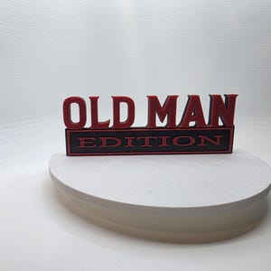 May include: A black and red sign that reads "OLD MAN EDITION". The letters "OLD MAN" are in red, raised lettering. The word "EDITION" is in red lettering on a black background.