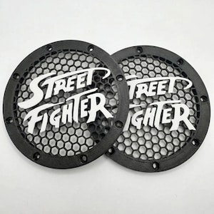 Può includere: Due griglie circolari nere per altoparlanti con un motivo a rete esagonale e il logo bianco Street Fighter centrato su ciascuna. Ogni griglia presenta un anello esterno solido con fori di montaggio distanziati uniformemente.