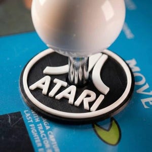 Könnte beinhalten: Ein weißer, kugelförmiger Arcade-Joystick-Griff auf einer schwarzen, runden Basis mit dem weißen Atari-Logo und Schriftzug. Der Joystick befindet sich auf einem blauen Bedienfeld, auf dem teilweise Text zu sehen ist, darunter MOVE und LAST FIGHTER.