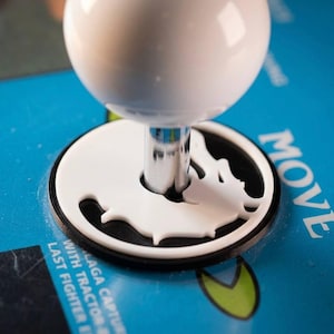 May include: A white spherical arcade joystick handle mounted on a chrome metal shaft, featuring a white circular dust cover with a dragon silhouette cutout, set against a blue control panel with text reading MOVE and LAST FIGHTER.