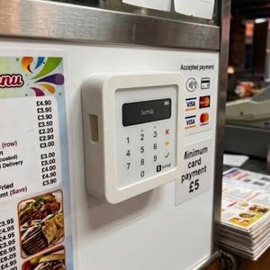 May include: A white SumUp card payment terminal mounted on a wall next to a menu. The device features a digital screen displaying the brand name, a numeric keypad, and symbols for card and contactless payments. Text on the wall indicates accepted payment methods.