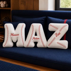 May include: Three white letter-shaped pillows spelling M, A, and Z. Each pillow features red baseball-style stitching patterns along the edges. The letters are personalized with the embroidered red names Mason, Aiden, and Zoe respectively.
