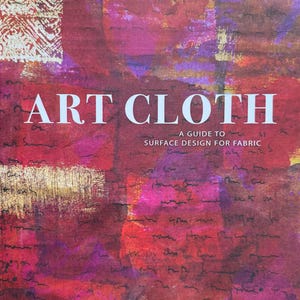 May include: A softcover book titled Art Cloth: A Guide to Surface Design for Fabric by Jane Dunnewold. The cover features an abstract background of layered red, magenta, and purple hues with gold accents and faint, handwritten script text.