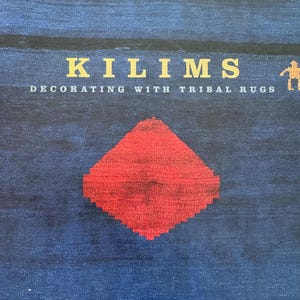 May include: A book cover with a deep blue textured background featuring a large red diamond shape in the centre. The text reads KILIMS DECORATING WITH TRIBAL RUGS and ELIZABETH HILLIARD. Small gold-coloured animal figures are at the top corners.