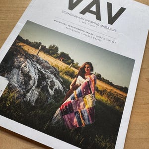 Peut inclure: Un exemplaire du magazine VÄV Scandinavian Weaving, n° 2, 2018. La couverture blanche présente un texte noir en gras et une photographie d'une personne dans un champ, drapée dans un textile tissé multicolore à motifs avec des franges.