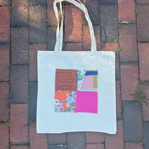 May include: A cream-colored canvas tote bag featuring a central square patch made of various fabric scraps. The patch includes sections of red floral print, orange and blue floral patterns, solid hot pink, and light blue geometric shapes.