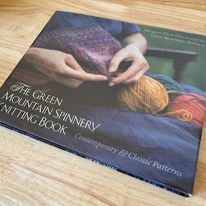 May include: A hardcover book titled The Green Mountain Spinnery Knitting Book: Contemporary & Classic Patterns by Margaret Klein Wilson and the Green Mountain Spinnery, featuring a cover image of hands knitting with colorful yarn.