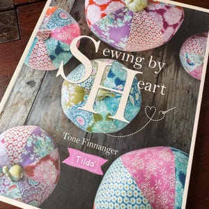May include: A softcover book titled Sewing by Heart by Tone Finnanger, featuring the Tilda brand logo. The cover displays several round, fabric-covered pumpkin-shaped ornaments made from various colorful floral and patterned cotton fabrics.