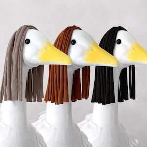 May include: Three white goose sculptures with yellow beaks and black eyes, each adorned with a different color of fabric hair: taupe, brown, and black. The sculptures are displayed against a plain white background.