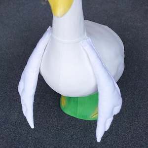 May include: A white goose head and body costume with a yellow beak and green base. The wings are white and extend outward. The costume is made of soft fabric.