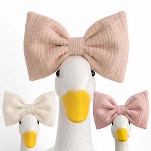 May include: Three white goose ornaments with yellow beaks and black eyes, each wearing a large, textured bow. The bows are in shades of pink and cream, creating a soft, whimsical look. The bows are made of a soft, textured fabric.