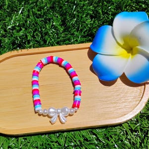 Puede incluir: Una colorida pulsera de cuentas con un dije de lazo blanco y detalles de perlas. La pulsera presenta cuentas rosas, azules y blancas. Descansa sobre una bandeja de madera junto a una flor azul y blanca, sobre un fondo verde.
