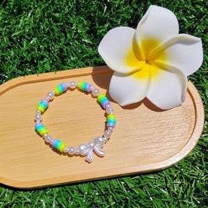 Puede incluir: Una pulsera hecha a mano con cuentas de colores arcoíris y pequeñas perlas blancas. La pulsera presenta un dije de lazo blanco. Se exhibe en una bandeja de madera con una flor blanca y amarilla.