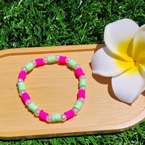 May include: A vibrant bracelet featuring pink and pale green beads, with small white beads between them. It rests on a wooden tray, beside a white flower with a yellow centre, against a background of green grass.