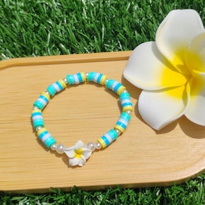 May include: A colorful bracelet with turquoise, yellow, and white beads, accented with a small white flower charm. The bracelet rests on a wooden tray next to a large white and yellow flower. The background is green.