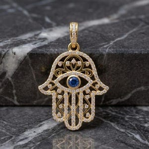 May include: Gold-toned Hamsa hand pendant encrusted with small, clear gemstones. The pendant features a central blue gemstone eye. The intricate design is set against a dark, marbled surface.
