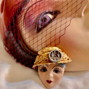 Könnte beinhalten: Ein Vintage-Gesicht aus Porzellan mit langen Wimpern, roten Lippen und einem kleinen Strohhut mit einem silbernen Knopf. Ein roter Netzschleier bedeckt den oberen Teil eines größeren Gesichts im Hintergrund.
