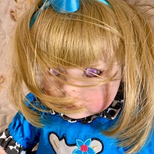 May include: A doll with blonde hair and purple eyes, wearing a blue dress with a cow graphic. The doll has a blue headband with a bow. The dress has black and white patterned sleeves and collar.