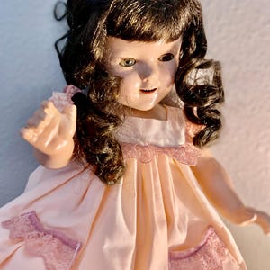 Könnte beinhalten: Eine Vintage-Puppe mit braunen Locken und grünen Augen. Die Puppe trägt ein hellrosa Kleid mit Spitzenbesatz und -akzenten. Das Gesicht der Puppe hat einen sanften Ausdruck, und die Gesamtästhetik erinnert an eine vergangene Ära.