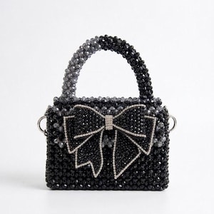 Puede incluir: Un bolso estructurado cubierto completamente por cuentas facetadas negras y gris oscuro. El bolso presenta un lazo central prominente hecho de cuentas negras con un borde de pedrería plateada. Incluye un asa de cuentas y anillos laterales de metal plateado.