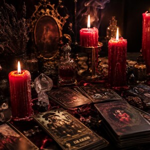 Op de afbeelding: Een verzameling tarotkaarten verspreid over een donker oppervlak, vergezeld door drie brandende rode stompkaarsen, kleine glazen flesjes, een decoratieve spiegel en gedroogde lavendel. De sc&egrave;ne wordt verlicht door warm kaarslicht.