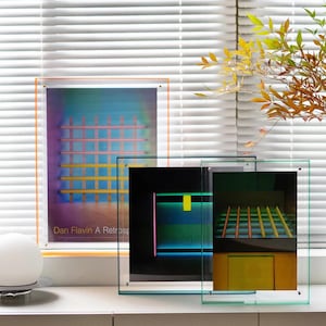 May include: Three rectangular acrylic frames holding art prints. The left frame has an orange border and displays a Dan Flavin A Retrospective poster with a grid pattern. The two right frames are clear, showcasing abstract neon light art prints.