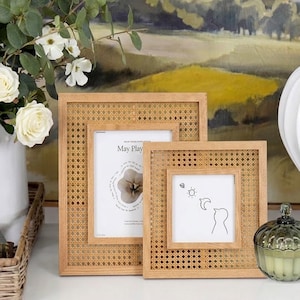May include: Two light-toned wooden picture frames featuring a woven cane rattan border. The larger rectangular frame holds a print with the text May Play, while the smaller square frame displays a line drawing of a sun, moon, and bird.