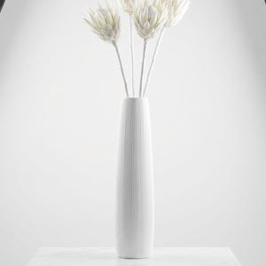 May include: A white, vertically-lined ceramic vase holds several white, spiky flower stems. The vase sits on a white, rectangular surface. The background is a bright white, with studio lighting visible on either side of the frame.