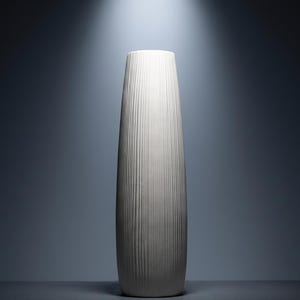May include: A tall, off-white ceramic vase with vertical ribbed detailing. The vase has a slender, elongated shape, wider at the base and tapering towards the top. The background is a gradient of dark blue to light blue, with a spotlight overhead.