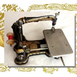 May include: A vintage black cast iron sewing machine featuring intricate gold floral scrollwork patterns, a silver-toned metal base plate, and a small wooden spool holder with a red thread bobbin attached to the side of the frame.