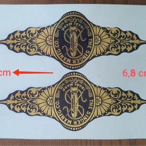 Könnte beinhalten: Zwei identische schwarze und goldene Aufkleber mit dem Singer Mfg. Co. Logo und filigranen Mustern. Die Aufkleber messen 18 cm in der Länge und 6,8 cm in der Höhe, wie durch die roten Pfeile und den Text auf dem weißen Blatt angegeben.