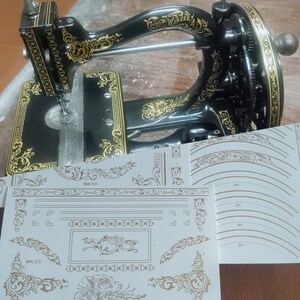 Può includere: Una macchina da cucire vintage nera con intricati motivi floreali dorati e il testo Wanzer A, accompagnata da fogli bianchi con bordi decorativi dorati e disegni angolari etichettati WA 1/3 e WA 2/3 per scopi di restauro.