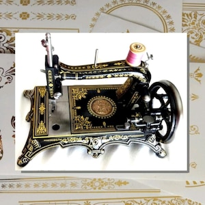 May include: A vintage-style black sewing machine featuring intricate gold filigree patterns across its frame. The machine includes a silver metal needle plate, a black handwheel on the right side, and a single spool of pink thread positioned on top.