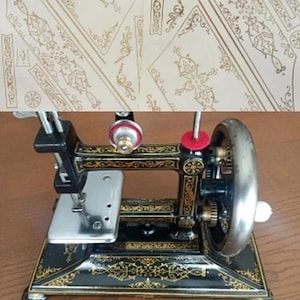 May include: A black metal sewing machine featuring intricate gold filigree patterns across its frame. The machine includes a silver-colored needle plate, a large side handwheel, and a red spool pin washer, showcasing a detailed vintage design.