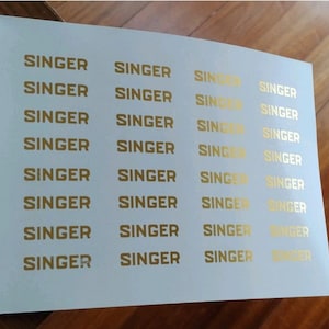 Pode incluir: Uma folha retangular branca apresentando uma grade de trinta e duas etiquetas individuais na cor dourada. Cada etiqueta exibe a palavra SINGER impressa em uma fonte em negrito, sem serifa e em letras maiúsculas.