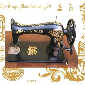 Op de afbeelding: Een zwarte vintage Singer naaimachine met gouden bloemmotieven en de tekst The Singer Manufacturing Co. en Fabrication Anglaise. De machine is voorzien van een groot handwiel, een naaldstang en een centraal goudkleurig logo op de basisplaat.