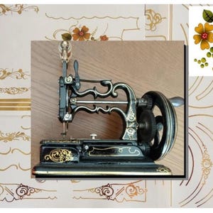 Peut inclure: Une machine à coudre manuelle noire vintage ornée de motifs dorés en volutes sur son cadre en métal. L'appareil comprend un volant latéral avec une manivelle et une barre à aiguille, sur un fond composé de décalcomanies florales dorées.