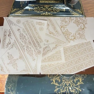 Puede incluir: Una máquina de coser vintage negra con patrones de filigrana dorados, acompañada de varias hojas de papel con diseños y bordes decorativos de volutas doradas, y un primer plano de la superficie negra de la máquina con detalles dorados.
